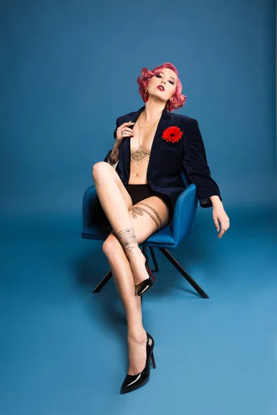 Sexy tattoed pin up girl in jacket with boutonniere posing on armchair infront of blue background — Stock Photo