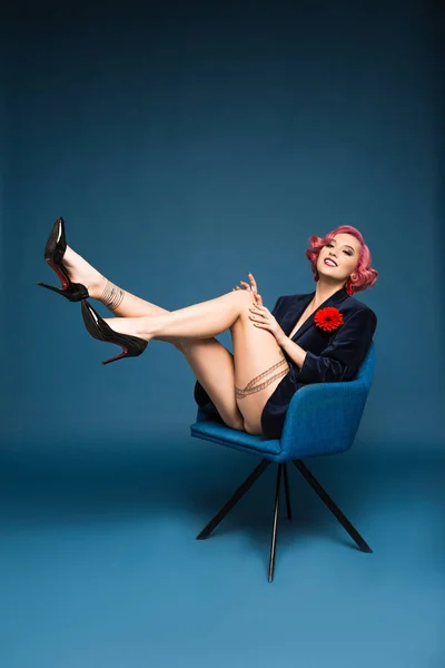 Beautiful tattoed pin up girl in jacket with boutonniere posing on armchair infront of blue background — Stock Photo