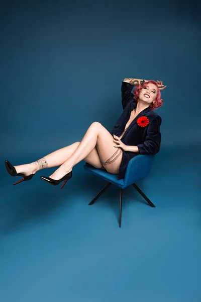 Belle tatouage pin up fille en veste avec boutonnière posant sur fauteuil devant fond bleu — Photo de stock