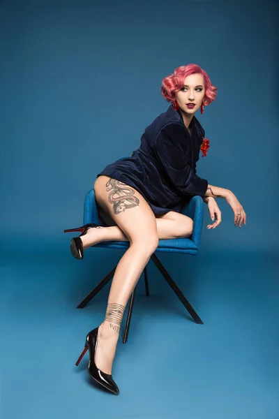 Sexy tattoed pin up girl in jacket posing on armchair infront of blue background — Stock Photo