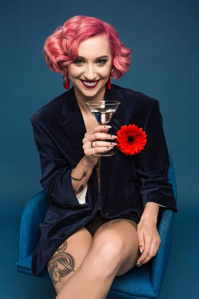 Красивая татуированная девушка в куртке с бутоньеркой и бокалом вина перед синим фоном — стоковое фото