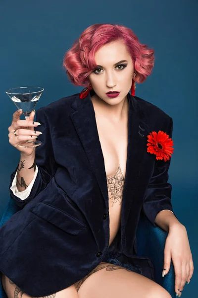 Beautiful tattoed pin up girl in jacket with boutonniere and wineglass infront of blue background — Stock Photo