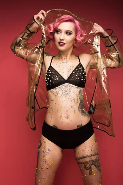 Sexy pin up fille avec des tatouages en lingerie et imperméable infront de fond rouge — Photo de stock