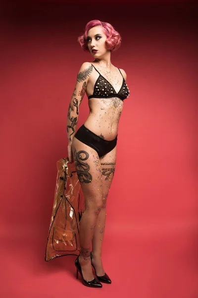 Attraente pin up ragazza con tatuaggi in lingerie tenendo impermeabile davanti a sfondo rosso — Foto stock