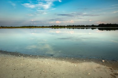Kum kıyısı ve sakin göl, ufukta ve gökyüzünde bulutlar