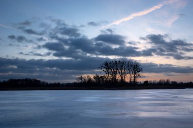 Donmuş göl, ufuktaki ağaçlar ve gün batımından sonra gökyüzünde kara bulutlar.