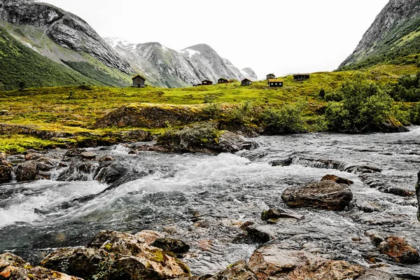 Bergdorf mit kleinen Häusern und Holzhütten mit Gras auf dem Dach in einem Tal. Die Hütten stehen auf grünem Gras und Weide. Berge sind mit Schnee bedeckt. — Stockfoto