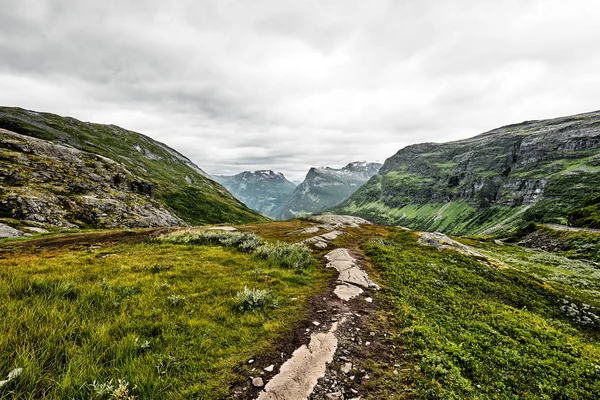 Weg über grüne Weiden in den Bergen Westnorwegens mit Schnee auf den Gipfeln und dunklem Himmel — Stockfoto