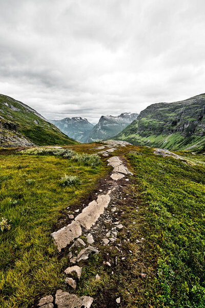 Path over green pasture in the mountains of Western Norway with snow on the summits and a dark cloudy sky