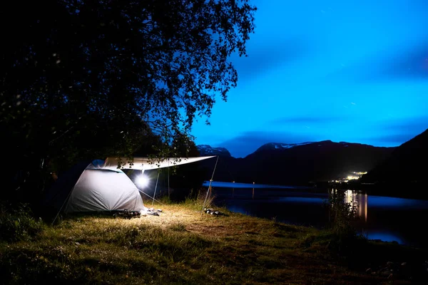 Bulutlu bir mavi gökyüzü ve yıldızlar ile gece boyunca plaj ve Norveç dağlarında fiyort ışıklı bir çadır kamp. 
