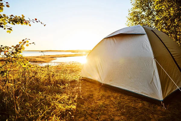 White tent standing at a beach camping spot at lake Vanern in Sweden. The sun is shining and soon will be sunset. Tent is standing on the right side covered by trees  and plants. Water reflections.