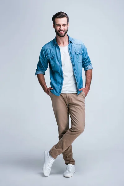 Modern ve rahat. Kameraya gülümse whilestanding beyaz arka plan ile seyir yakışıklı genç adam tam uzunlukta