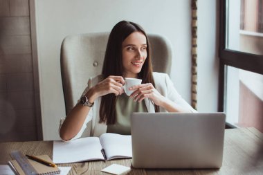 Kahve molası. Kahve fincanı tutarak ve ofis masasında otururken gülümseme ile pencereden bakıyor güzel genç iş kadını