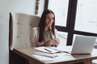 Büyük iş mesaj. Akıllı telefonunu kullanarak ve ofis masasında otururken gülümseme ile kameraya bakarak güzel genç iş kadını