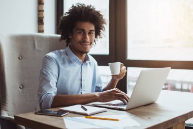 İyi günler iş başında. Diz üstü kullanarak ve ofis masasında otururken gülümseme ile kameraya bakarak yakışıklı genç işadamı yan görünüm