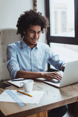 Onun çalışma günü zevk. Yakışıklı genç işadamı ofis masasında otururken gülümseme ile dizüstü bilgisayar kullanarak