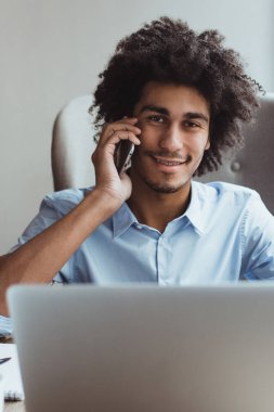 Merhaba,. Cep telefonu üzerinde konuşurken ve ofis masasında otururken gülümseme ile kameraya bakarak yakışıklı genç işadamı