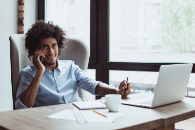 Neşeli işadamı iş başında. Cep telefonu üzerinde konuşurken ve ofis masasında otururken gülümseme ile kameraya bakarak yakışıklı genç işadamı