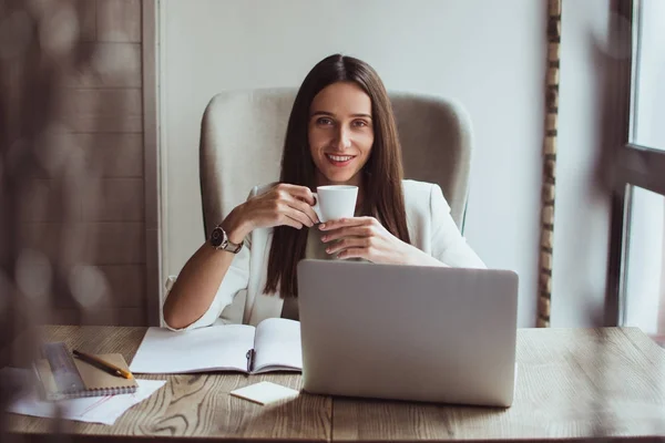 What can be better then fresh cup of coffee? Beautiful young businesswoman holding coffee cup and looking at camera with smile while sitting at her office desk