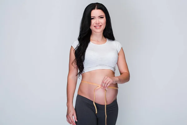 We grow so fast! Beautiful young pregnant woman looking at camera with smile and holding measure tape around her belly while standing at white background