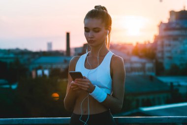 Koşu için onun favori listesi. Spor giyim sanayi kenti görünümü karşı dururken onun smartphone ile gündoğumu kullanarak genç güzel kadın