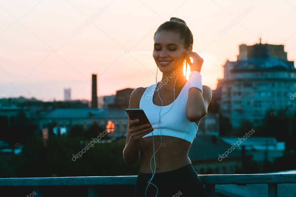 In love with her city. Young beautiful woman in sportswear using her smartphone with smile while standing against industrial city view with sunrise