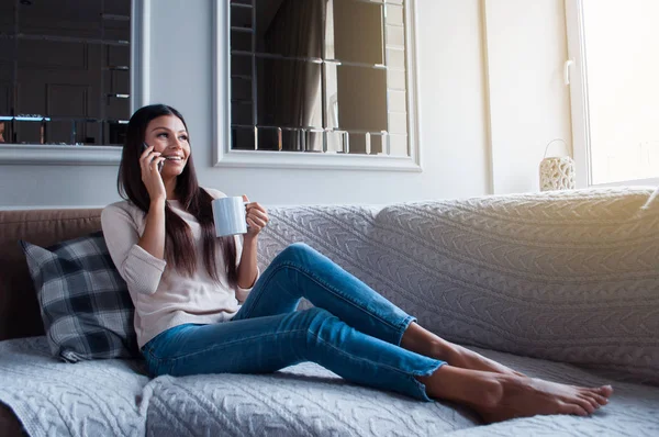 Have you heard latest news? Full length of beautiful young woman holding coffee mug and talking on mobile phone with smile while sitting on the sofa in home interior