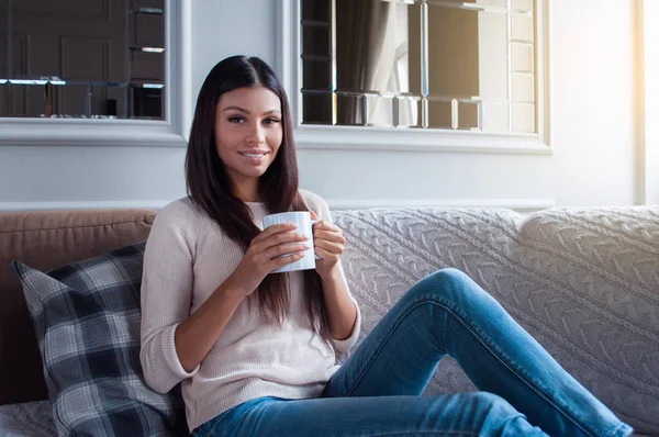 Spending perfect morning at home. Beautiful young woman holding coffee mug and looking at camera with smile while sitting on the sofa in home interior