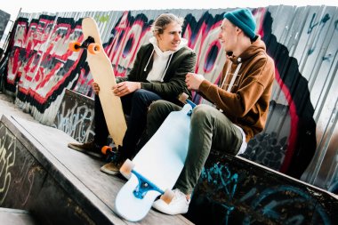 Kaykay tutkudur. İki genç erkek birbirinizle longboards açık havada kaykay parkı içinde otururken gülümseme ile konuşmak 