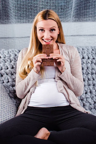 Guilty pleasure. Beautiful young pregnant woman eating chocolate and looking at camera with smile while sitting on the sofa in home interior