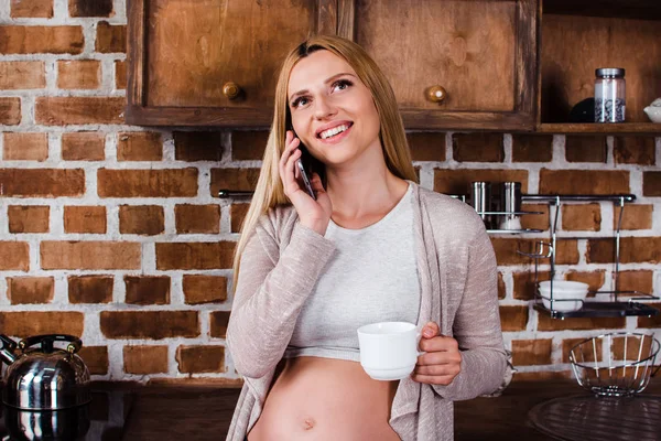 So nice to hear you! Beautiful young pregnant woman talking on phone and looking away with smile while standing at kitchen interior
