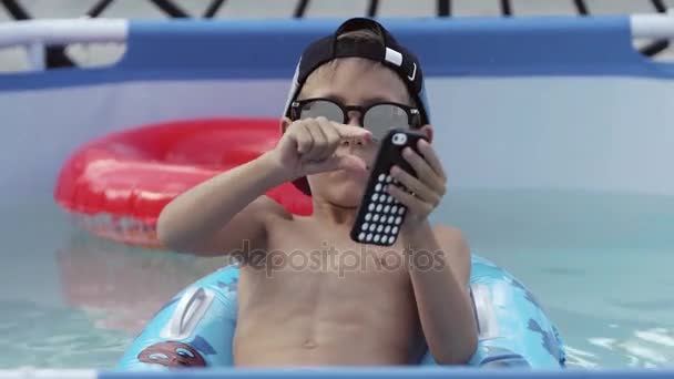 O menino está tocando o telefone e relaxando na piscina — Vídeo de Stock