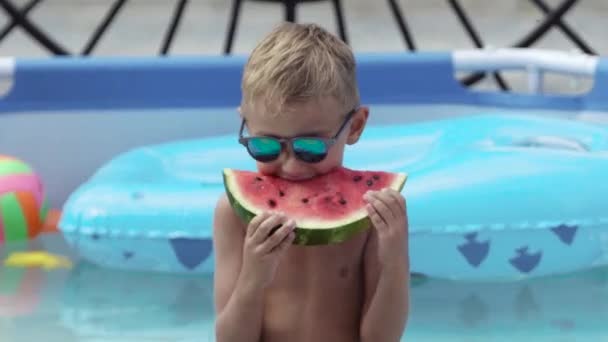 Niño en gafas negras come sandía en la piscina — Vídeos de Stock