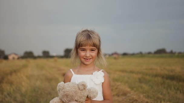 Menina bonita com cabelos longos em um vestido branco caminha campo com um urso de brinquedo macio — Vídeo de Stock