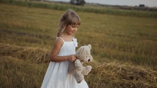 Маленькая девочка с красивыми темными глазами ходит по полю после дождя и играет игрушечный медведь — стоковое видео