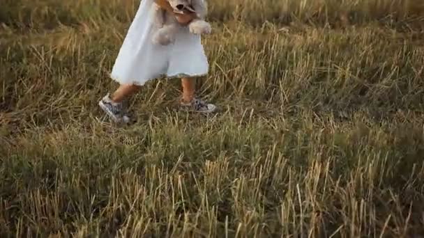 Een meisjes schoenen in een kitty en gekleed in een witte jurk wandelingen op het veld en wordt gespeeld door een beer speelgoed — Stockvideo