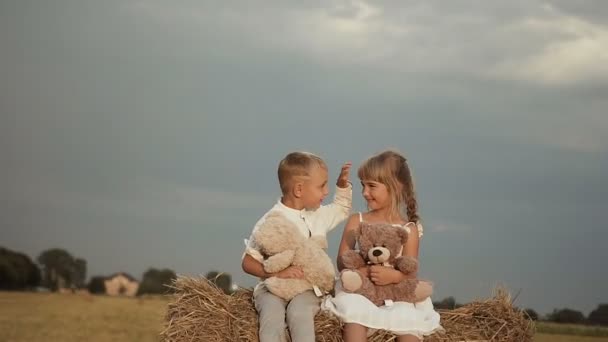 Happy children after the rain sit high on straw and play toy bears. A smiling boy straightens the girl hair — Stock Video