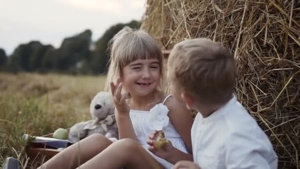 En liten flicka i en vit klänning vaknar en pojke som äter ett plommon. Barn i fältet sitta under höbalar — Stockvideo