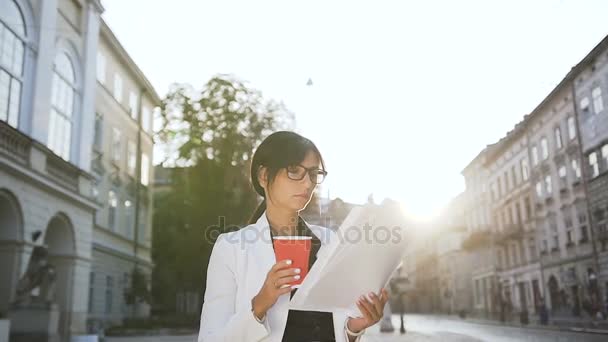 Una joven mujer de negocios con gafas paseando por la ciudad y tomando café mientras mira los documentos — Vídeos de Stock