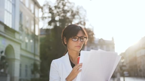 Una joven mujer de negocios con gafas paseando por la ciudad y tomando café mientras mira los documentos — Vídeo de stock