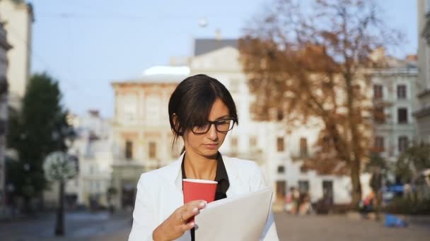 Una giovane donna d'affari con gli occhiali che cammina per la città e beve caffè guardando i documenti — Video Stock