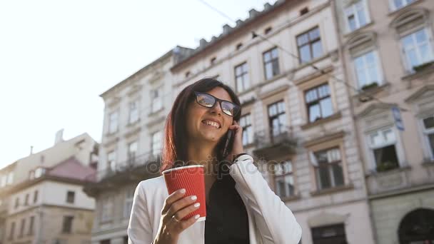 Una joven mujer de negocios caminando por la ciudad, hablando por teléfono y tomando café — Vídeos de Stock