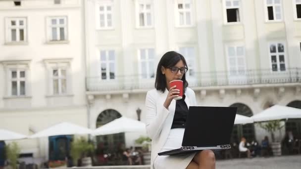 Una joven mujer de negocios con gafas trabaja en un ordenador de la ciudad y bebe café — Vídeos de Stock