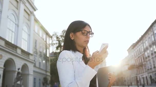 A young business woman wearing glasses in the morning goes to hte city and drops sms — Stock Video