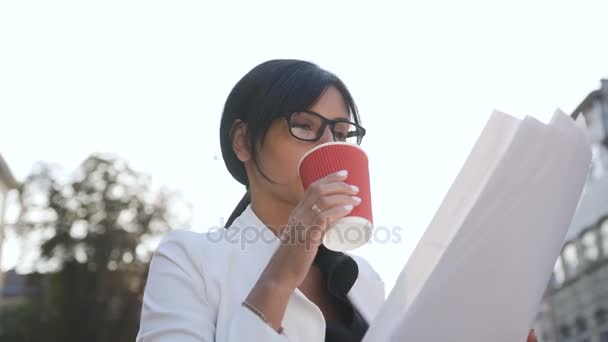 Een jonge vrouw die een bril gaat aan het werk in de ochtend, drinkt koffie en onderzoekt documenten — Stockvideo