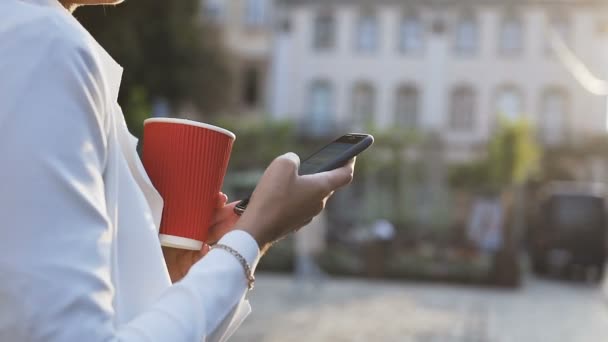 Молода жінка гуляє містом, п'є каву і пише повідомлення по телефону — стокове відео