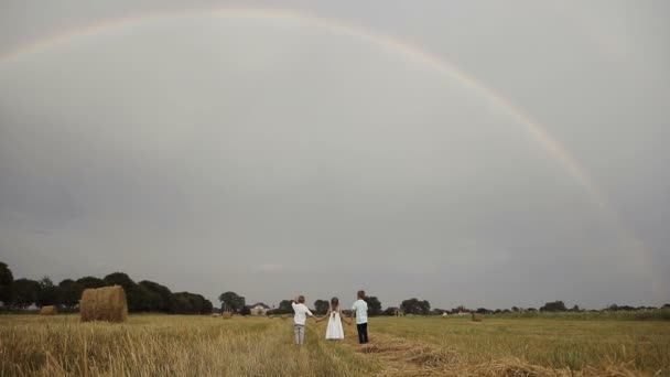 Una niña y dos niños corren un campo amarillo con palos de paja para encontrarse con el arco iris — Vídeos de Stock