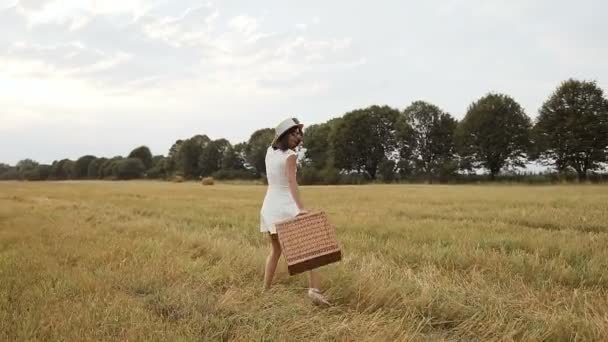 Ένα νεαρό κορίτσι που φοράει ένα άσπρο φόρεμα με ένα ψάθινο καπέλο και μια βαλίτσα. Μπάλες άχυρο και άχυρο. Ηλιοβασίλεμα — Αρχείο Βίντεο