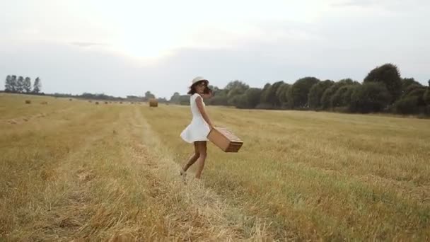 Mladá dívka, která nosí bílé šaty s slaměný klobouk a kufr. Sláma a slaměné balíky. Západ slunce — Stock video