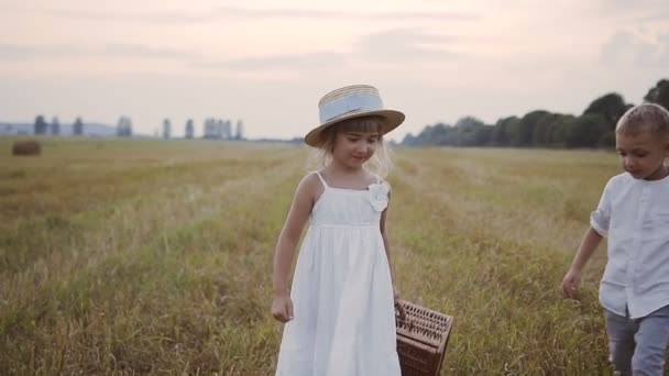 Un niño pequeño ayuda a una niña a llevar una maleta de paja en un vestido blanco. Los niños caminan en el campo. Puesta de sol — Vídeos de Stock